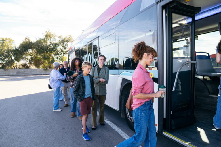 Mehrere Personen stehen in einer Schlange vor einem Elektrobus am Langzeitparken Warnemünde , während eine Frau mit Kaffeebecher einsteigt.