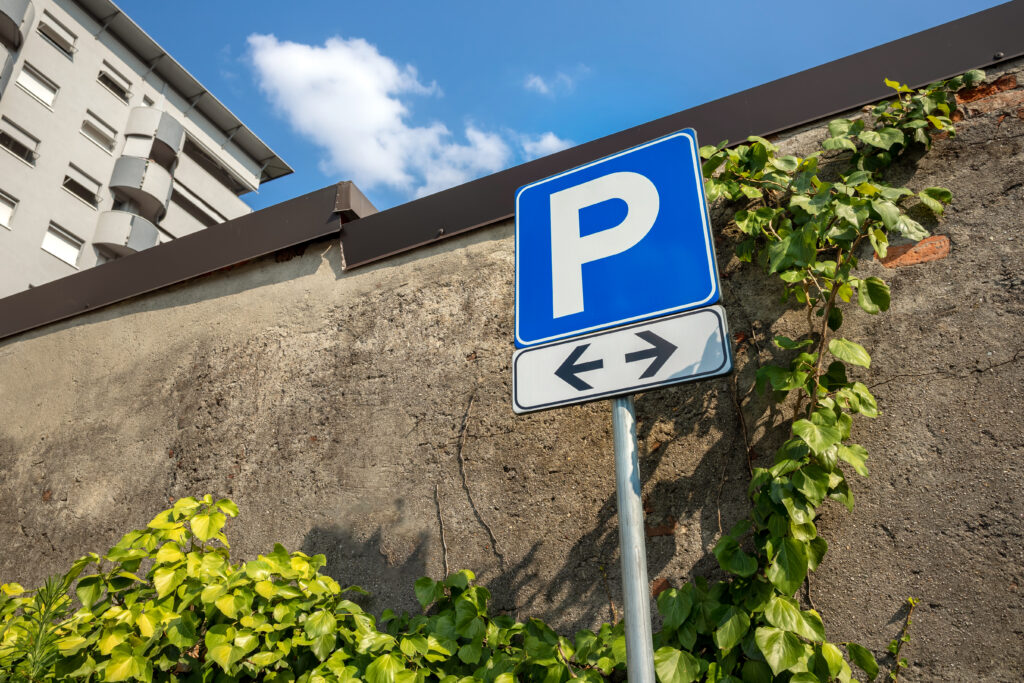 Parkschild mit Richtungspfeilen steht vor einer Mauer, an der Efeu emporwächst.