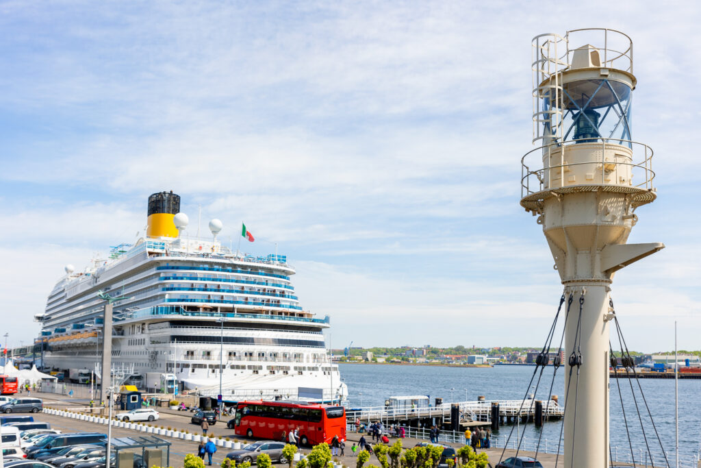 Großes modernes Kreuzfahrtschiff liegt am Hafen neben einem Leuchtturm und mehreren geparkten Autos